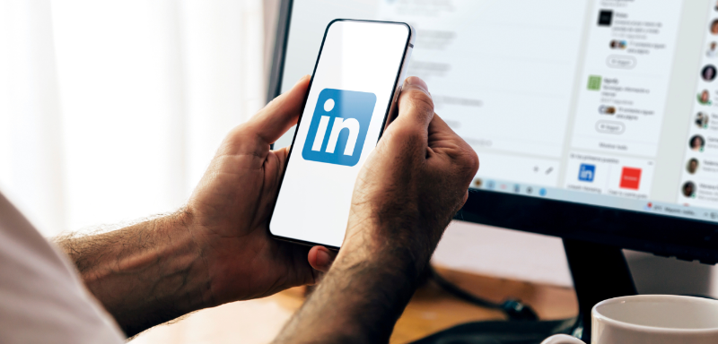 Logo do LinkedIn em celular, segurado por homem em frente a computador.